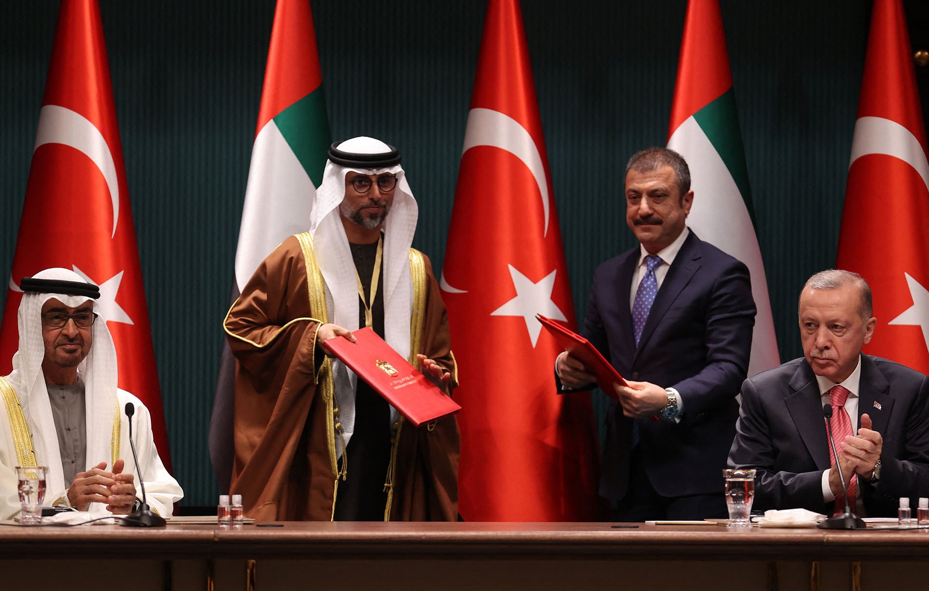 L'immagine mostra una cerimonia ufficiale con quattro uomini in abiti formali. Sullo sfondo si vedono le bandiere degli Emirati Arabi Uniti e della Turchia. Due degli uomini sono in piedi e sembrano scambiarsi documenti, mentre gli altri due sono seduti. L'atmosfera suggerisce un momento importante, probabilmente relativo a un accordo o a una collaborazione tra i due paesi.