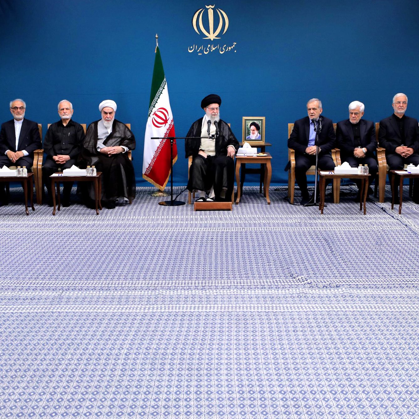 الصورة تظهر مجموعة من الأشخاص يجلسون في صفين. في الخلفية يوجد علم إيران، وهناك صورة للمرشد الأعلى. الأفراد يرتدون ملابس رسمية، ويبدو أنهم في مناسبة أو اجتماع رسمي. الغرفة تتميز بجو رسمي ومهيب، مع سجادة تغطي الأرض.
