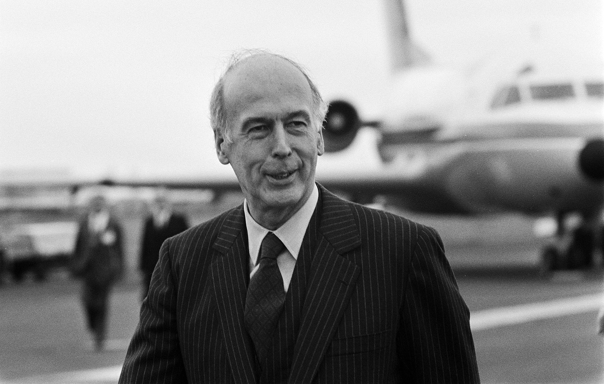 L'image montre un homme en costume rayé, marchant sur une piste d'aéroport. Il affiche un léger sourire et semble détendu. En arrière-plan, on peut apercevoir un avion et d'autres personnes qui se déplacent. L'ambiance générale est celle d'un moment de rencontre ou d'accueil, et la photographie est en noir et blanc, ce qui lui donne un caractère classique.
