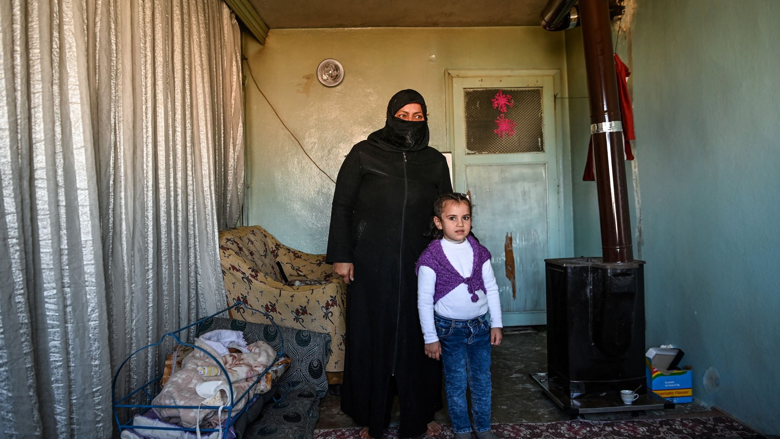 L'image montre une femme portant une tenue noire et un voile, se tenant à côté d'une petite fille. Elles sont à l'intérieur d'une pièce qui semble modeste, avec des murs verts pâles et un tapis au sol. À gauche, il y a une couchette avec un bébé endormi. Un poêle en métal est visible dans un coin de la pièce, et des rideaux en tissu léger sont tirés. L'ambiance est familiale, mais la simplicité du décor reflète une certaine précarité.