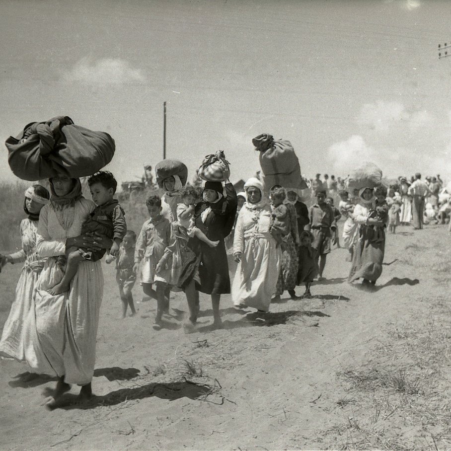 L'image montre un groupe de personnes marchant le long d'un chemin poussiéreux. Certaines femmes portent de lourdes charges sur la tête, tandis que d'autres tiennent des enfants par la main. La scène semble illustrer un moment de déplacement ou de migration, avec des paysages arides en arrière-plan. Des nuages blancs ponctuent le ciel, et on peut voir des lignes électriques au loin. L'atmosphère de la photo évoque la lutte et la détermination des personnes en marche.