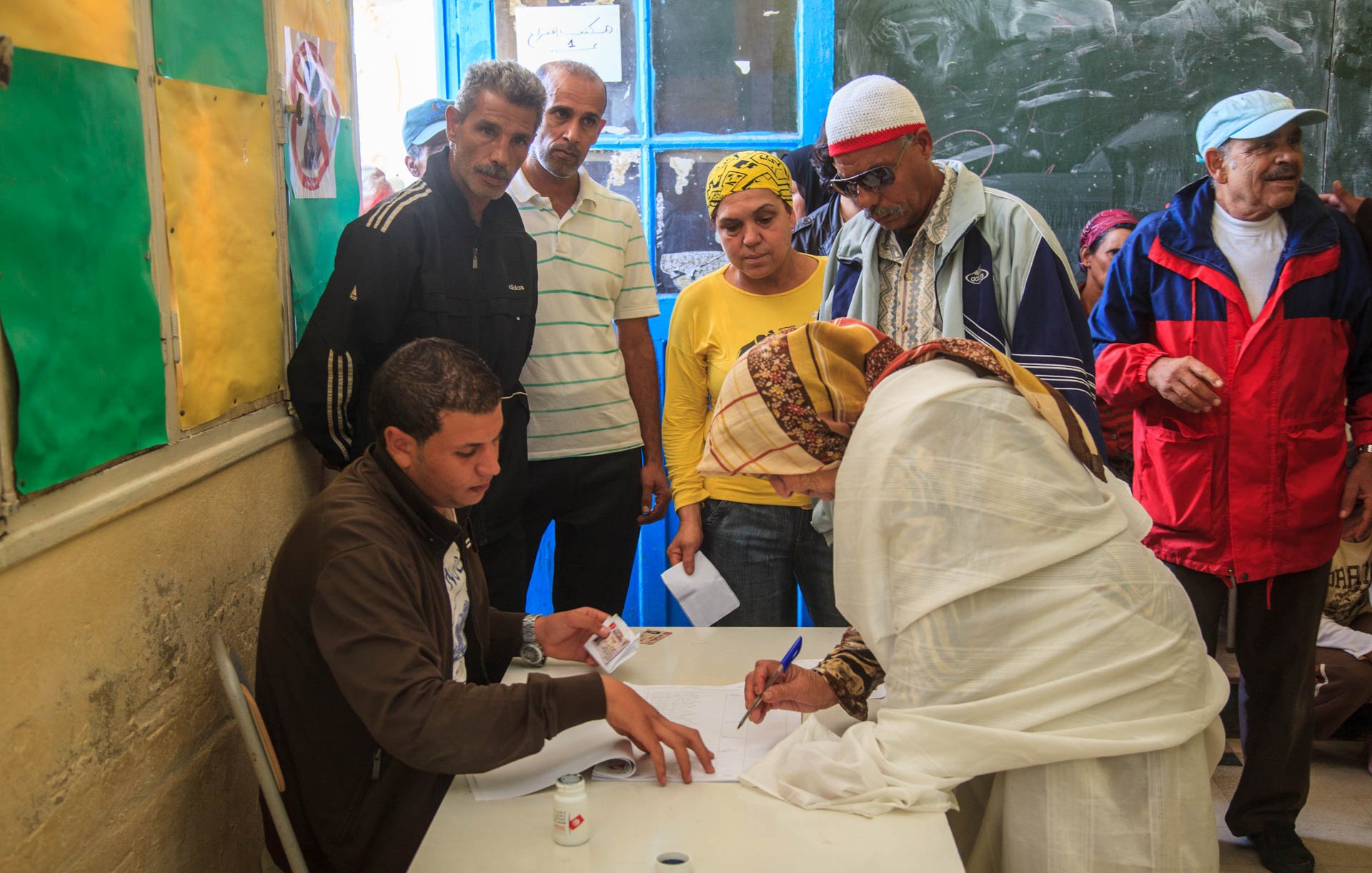 L'image montre un groupe de personnes se rassemblant autour d'un bureau dans une salle. Un homme est assis à une table, où il semble aider une femme, qui est en train de signer un document. Elle porte une tenue claire et un foulard. D'autres personnes, dont des hommes et des femmes, observent la scène, manifestant un intérêt. Les murs sont décorés de couleurs vives, et une fenêtre ouverte laisse entrer la lumière. L'environnement semble informer d'une activité communautaire, peut-être liée à un vote ou à une inscription.