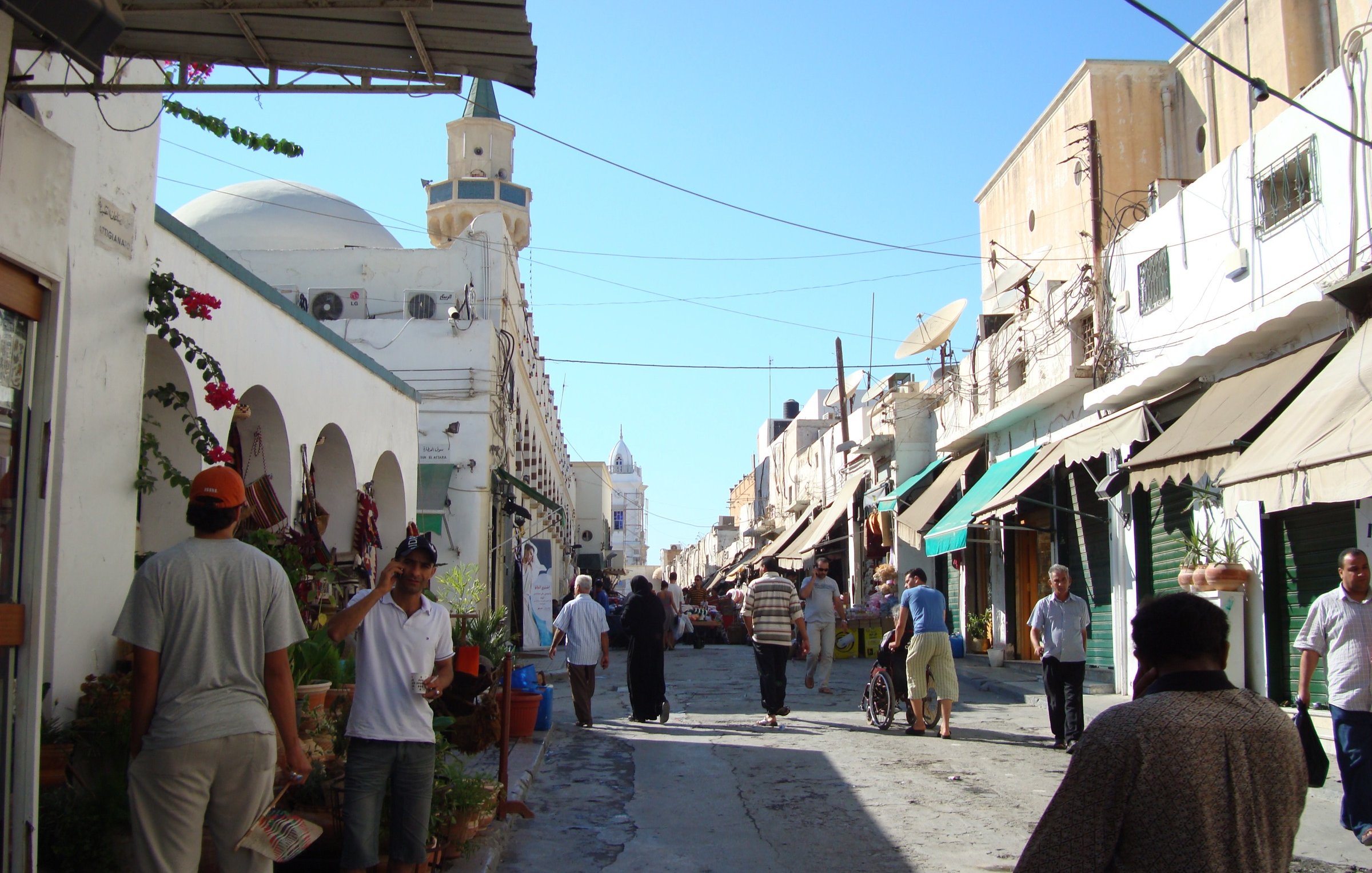 L'image montre une rue animée d'une ville typique, avec des bâtiments de style méditerranéen. On peut voir des personnes marchant le long de la rue, parmi lesquelles des hommes habillés en vêtements traditionnels. Des pots de fleurs et des végétaux décorent l'entrée de certains bâtiments, tandis que des ombrelles ou des auvents semblent offrir de l'ombre aux passants. Le ciel est dégagé et ensoleillé, créant une ambiance agréable.