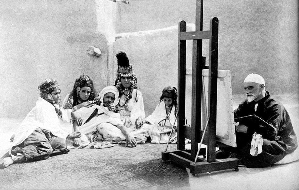 L'image montre un groupe de personnes assises autour d'un homme qui peint sur un chevalet. Les membres du groupe portent des vêtements traditionnels, certains avec des bijoux et des motifs distinctifs. L'homme qui peint semble concentré sur son travail, tandis que les autres l'observent attentivement. L'environnement paraît simple, avec des murs en argile en arrière-plan, ce qui donne un aperçu d'une ambiance culturelle et artistique.