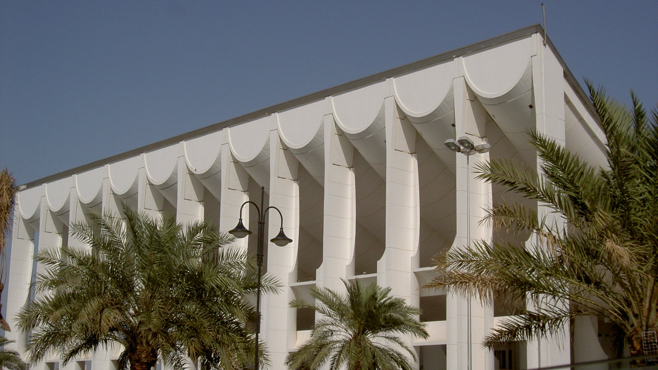 الصورة تظهر مبنى معماري متميز يتميز بتصميمه العصري. يتكون المبنى من واجهة بيضاء تحتوي على عموديات بارزة، مع استخدام الأقمشة البيضاء التي تزين السقف وتضفي عليه مظهراً فريداً. تحيط بالمبنى أشجار النخيل والحدائق الخضراء، مما يضفي جوًا منعشًا وجميلًا. السماء زرقاء صافية مما يعكس جمال اليوم.