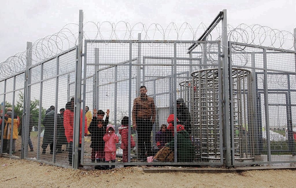 L'image montre un groupe de personnes à l'intérieur d'une structure grillagée, entourée de barbelés. On peut voir des adultes et des enfants, certains portant des vêtements colorés, semblant attendre dans un espace très restreint. L'ambiance semble préoccupante, suggérant une situation de confinement ou d'attente difficile. Le ciel est nuageux, ce qui contribue à une impression de morosité.
