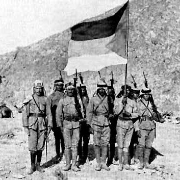 L'image représente un groupe de soldats posant devant un drapeau. Ils sont vêtus de uniforme militaire, portant des capes et des casques. En arrière-plan, on aperçoit un paysage montagneux et quelques tentes, suggérant un camp militaire. Les soldats, tenant leurs fusils, semblent être en position de parade ou de cérémonie. L'atmosphère de la photo évoque une période historique de conflit ou de guerre.