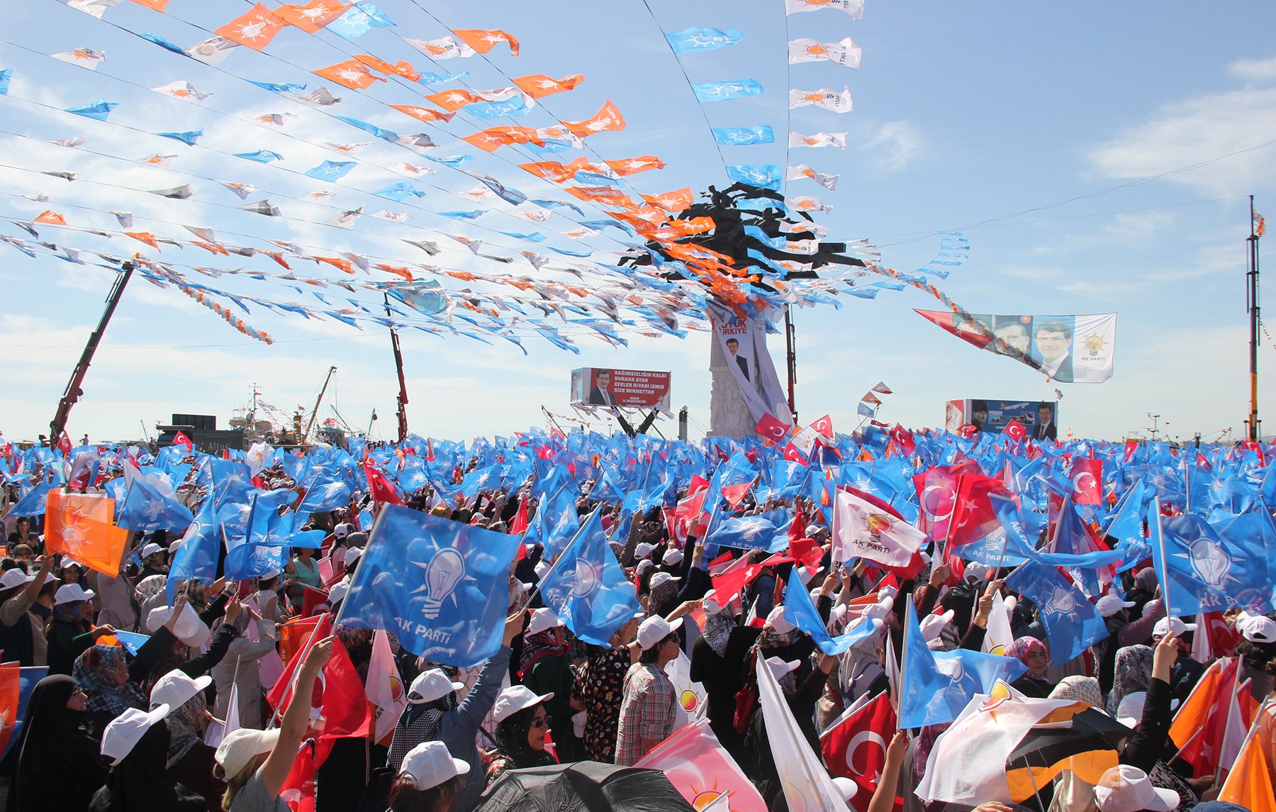 L'image montre une grande foule rassemblée lors d'un événement, probablement un rassemblement politique ou une manifestation. Des drapeaux de différentes couleurs, notamment bleu et orange, flottent dans l'air, créant une ambiance festive. Les participants portent des chapeaux blancs et semblent très engagés dans l'événement. Le ciel est clair, ce qui ajoute à l'énergie de la scène. En arrière-plan, des affiches et des structures indiquent un cadre organisé pour cet événement public.