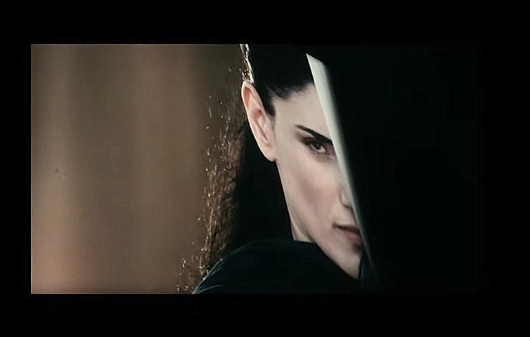 L'image montre une femme avec une expression intense et déterminée. Elle est à moitié cachée derrière un objet, ce qui accentue son regard perçant. Ses cheveux sont longs et foncés, et son attitude suggère qu'elle est dans une situation de confrontation ou de défense. L'éclairage met en avant son visage et crée une ambiance mystérieuse.