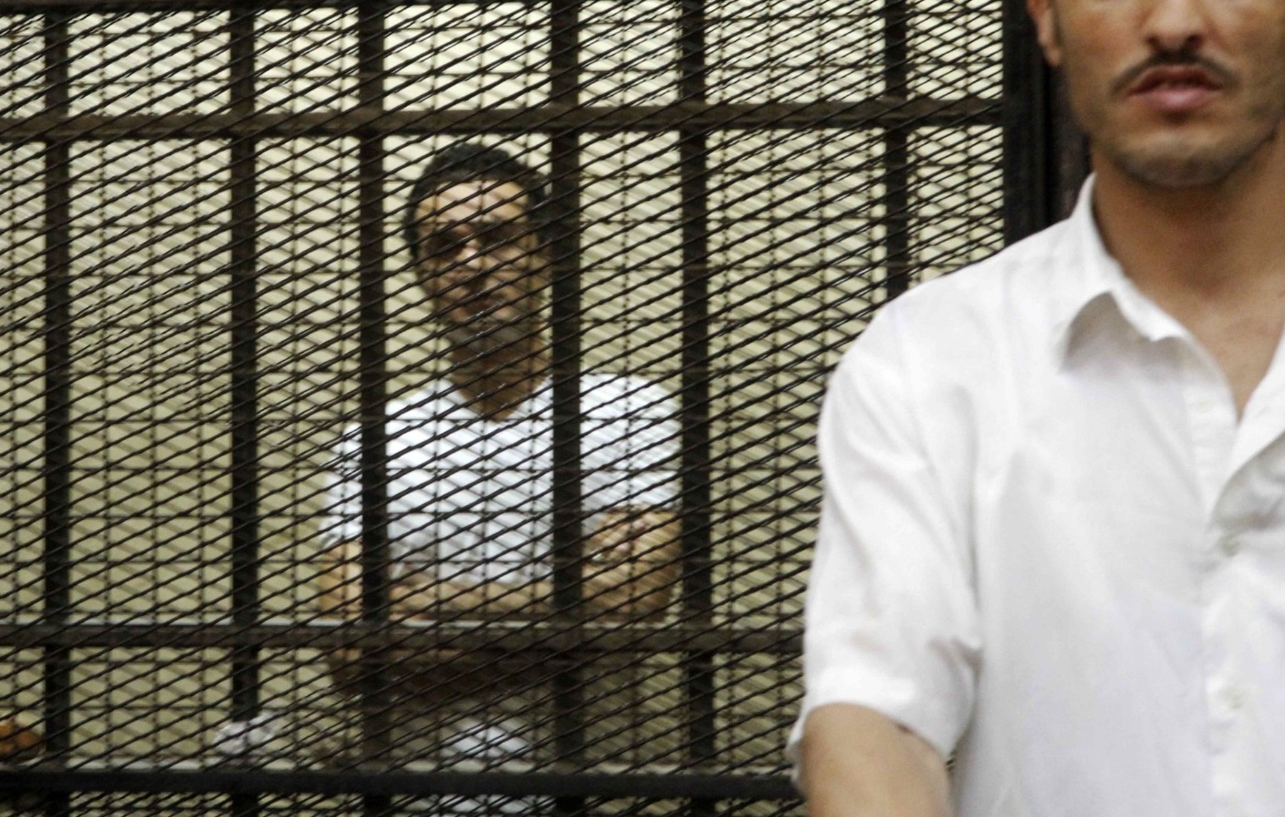 Deux hommes se tiennent derrière des barreaux, l'un visible et l'autre en arrière-plan.