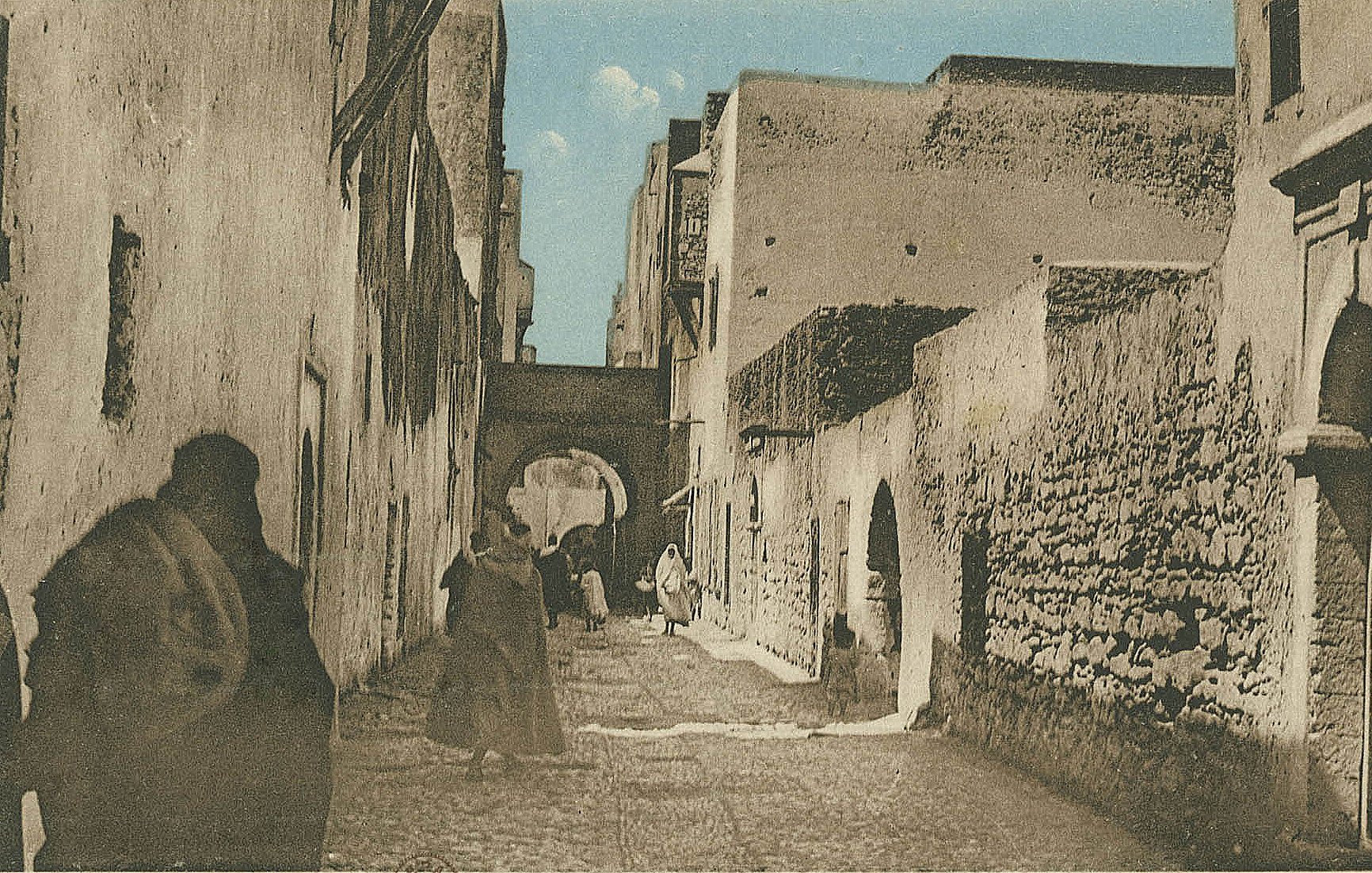 L'image représente une ruelle étroite et pavée, probablement dans une ville historique. Les murs des bâtiments sont en pierre, avec des textures vieilles et usées, suggérant un certain âge. On peut apercevoir des silhouettes de personnes marchant, certaines vêtues de longues robes traditionnelles. À l'arrière-plan, un arc architectural est visible, ajoutant une touche d'authenticité à l'environnement. Le ciel est dégagé, apportant une lumière douce à la scène. L'atmosphère semble paisible et évoque un lieu chargé d'histoire.