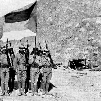 L'image représente un groupe de soldats posant devant un drapeau. Ils sont vêtus de uniforme militaire, portant des capes et des casques. En arrière-plan, on aperçoit un paysage montagneux et quelques tentes, suggérant un camp militaire. Les soldats, tenant leurs fusils, semblent être en position de parade ou de cérémonie. L'atmosphère de la photo évoque une période historique de conflit ou de guerre.