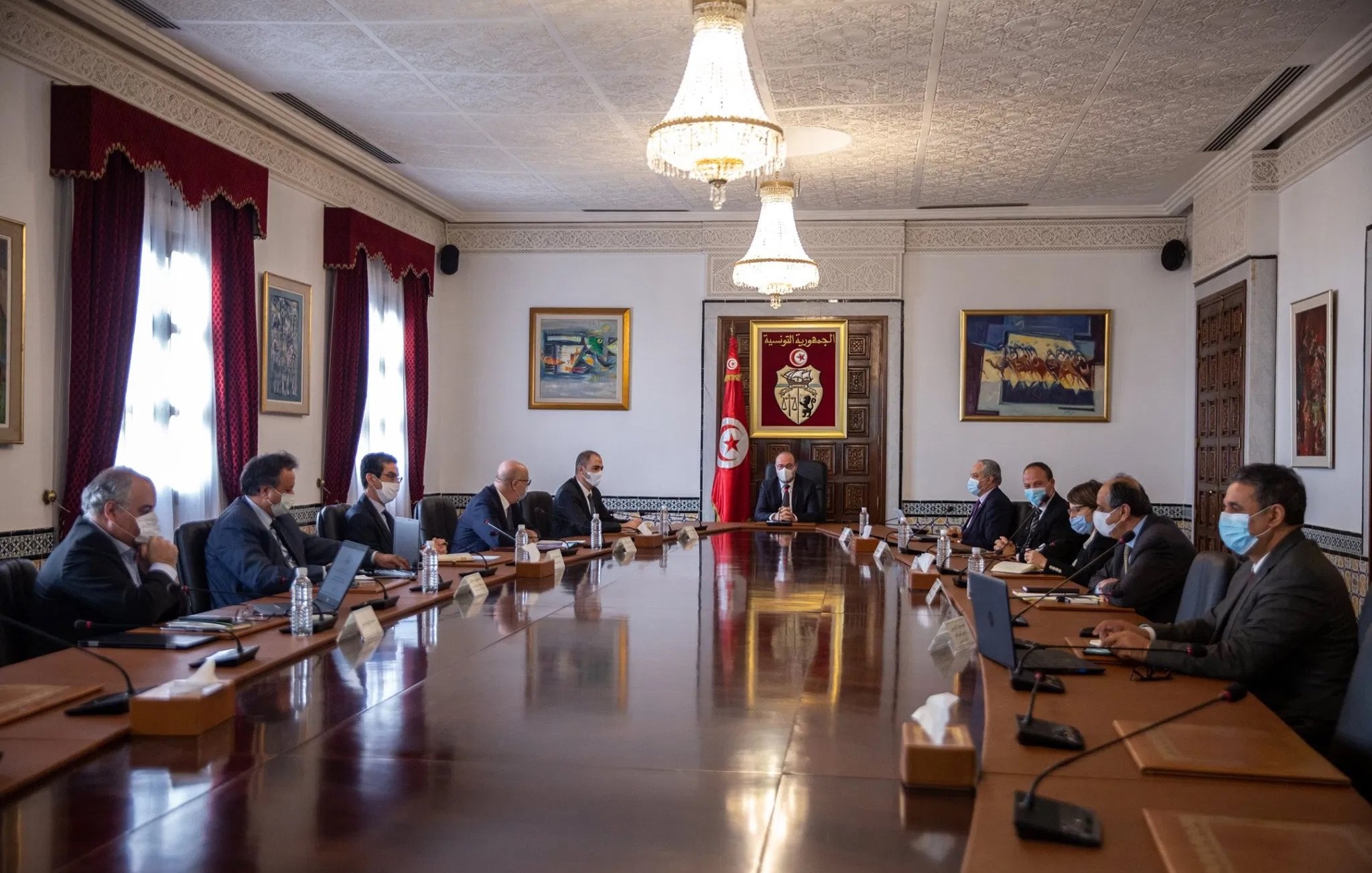 L'image montre une réunion qui se déroule dans une salle de conférence élégante. Il y a plusieurs personnes assises autour d'une grande table en bois. Au centre, un homme semble diriger la réunion, tandis que les autres participants l'écoutent attentivement. La salle est décorée de tableaux et présente des fenêtres avec des rideaux rouges. Tous les participants portent des masques, ce qui suggère une prudence sanitaire. L'atmosphère est formelle, typique d'une réunion gouvernementale ou d'affaires.