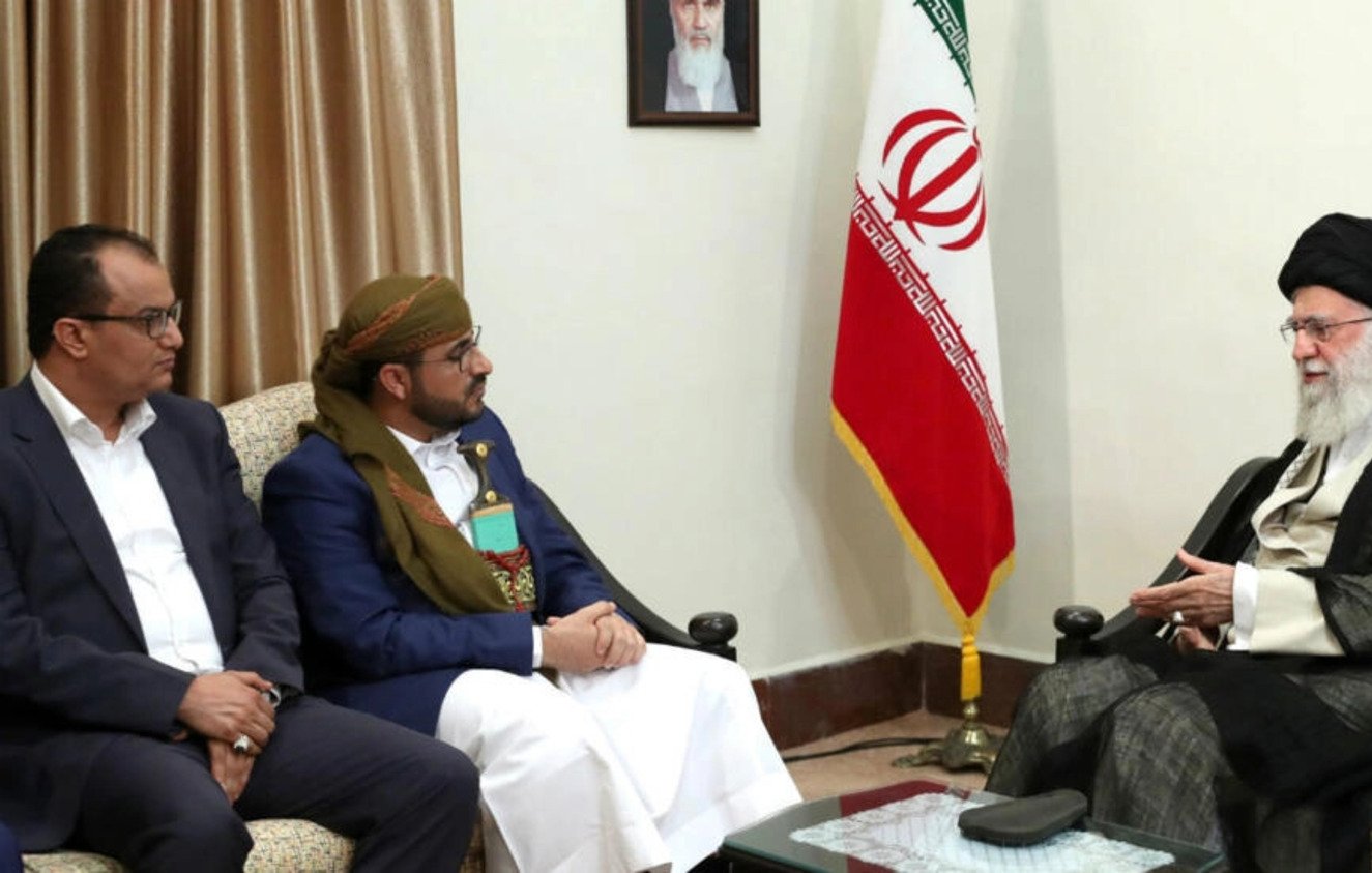 در این تصویر، دو مرد در حال گفتگو با یک شخصیت دیگر هستند. آن‌ها در یک مکان رسمی نشسته‌اند و تصاویر و پرچم‌هایی در پس‌زمینه دیده می‌شود. یکی از مردان لباس سنتی پوشیده و دیگری لباس رسمی دارد. شخصیت دیگر که صحبت می‌کند، عمامه و لباس سنتی ایران را به تن دارد. فضای گفتگو نشان‌دهنده یک جلسه ملاقات رسمی است.