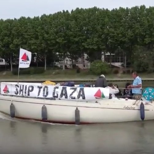 L'image montre un bateau naviguant sur une étendue d'eau. Sur le bateau, il y a des personnes présentes, et une banderole est accrochée, indiquant "SHIP TO GAZA". Le bateau arbore également plusieurs drapeaux, dont ceux de la Palestine et de la Suède, ainsi que d'autres drapeaux qui pourraient représenter différentes organisations ou pays. En arrière-plan, des arbres et des bâtiments sont visibles, suggérant un environnement urbain proche d'une rivière ou d'un port.