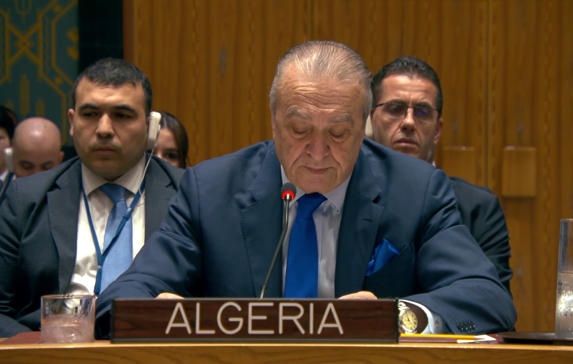 رجل يتحدث خلف منصة، يحمل لافتة الجزائر، مع أشخاص يجلسون خلفه في قاعة.