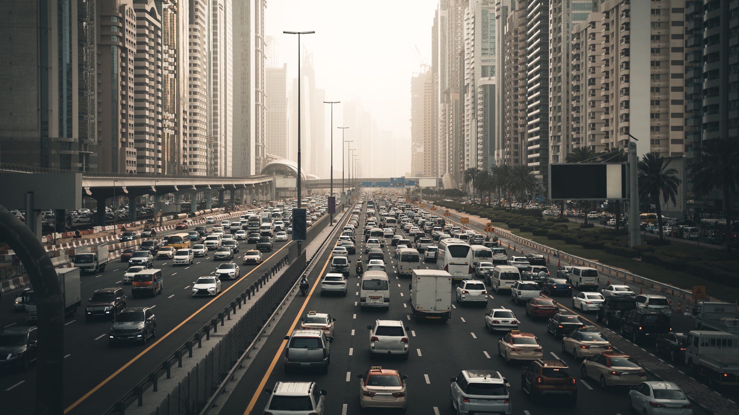 L'image montre une vue d'une route très fréquentée dans une grande ville moderne, entourée de gratte-ciel. On peut voir de nombreux véhicules qui circulent dans les deux sens, créant un embouteillage. La lumière du soleil est tamisée, donnant une atmosphère légèrement brumeuse, peut-être due à la pollution ou à la météo. Les bâtiments en arrière-plan sont imposants et reflètent l'architecture contemporaine, avec des façades en verre et en béton. Des palmiers sont visibles sur le côté de la route, ajoutant une touche de verdure à ce paysage urbain.