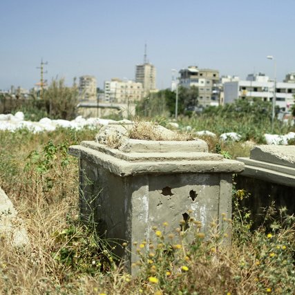 L'image montre un cimetière abandonné ou négligé, où des tombes en béton sont entourées d'herbes hautes et de végétation. On peut apercevoir, au fond, des bâtiments de plusieurs étages et des structures urbaines, ce qui indique une proximité avec une zone habitée. L'atmosphère est calme, mais on ressent un contraste entre l'état de mise à l'écart des tombes et l'activité de la ville environnante.