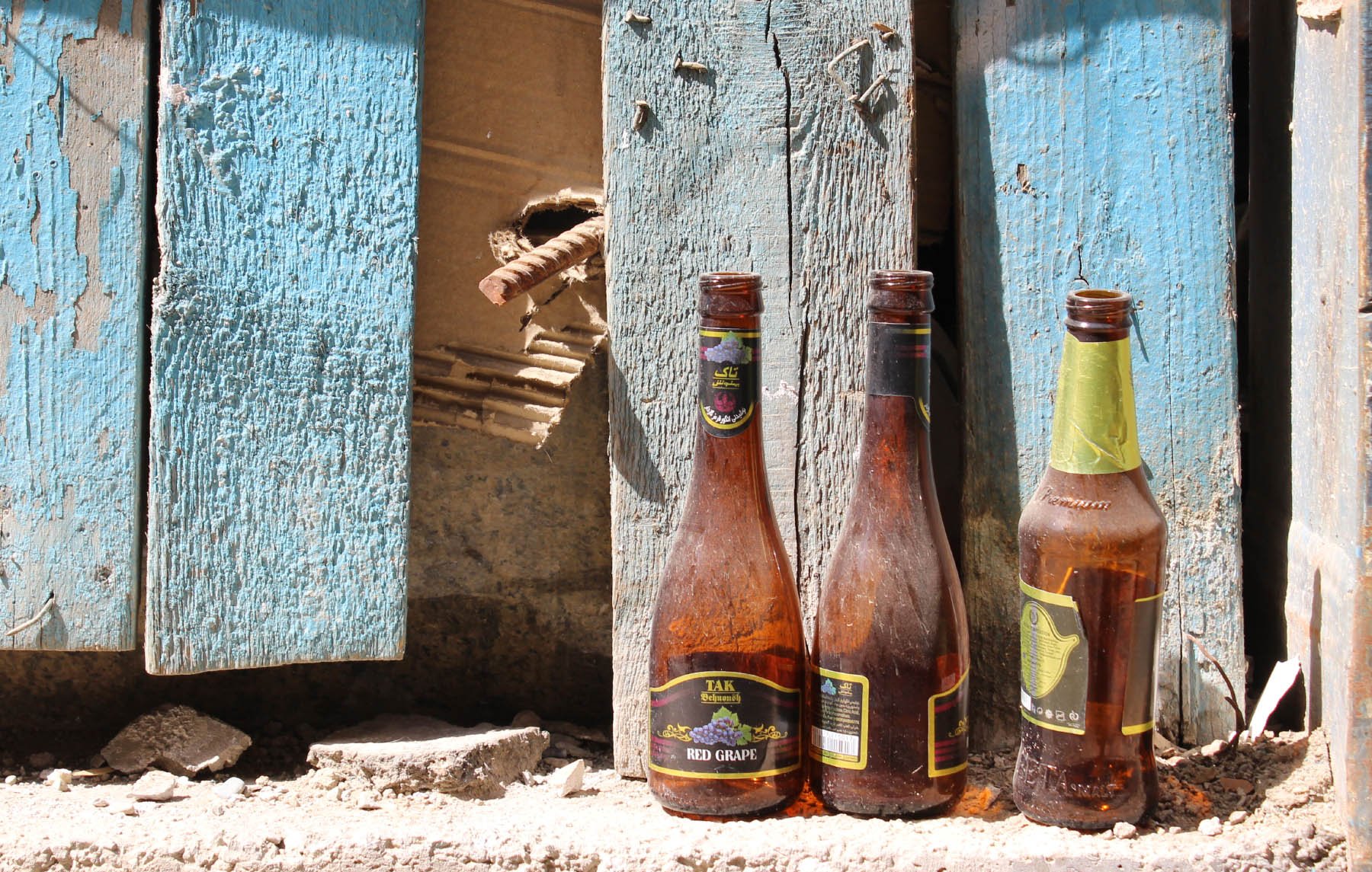 L'image montre trois bouteilles en verre vides, posées sur un sol poussiéreux. Elles sont de formes et de tailles différentes, et montrent des traces de saleté et d'usure. En arrière-plan, on aperçoit des planches de bois bleuies, dont certaines semblent endommagées, avec un trou visible. L'ensemble dégage une impression de désordre et de négligence, suggérant un espace abandonné ou en déclin.
