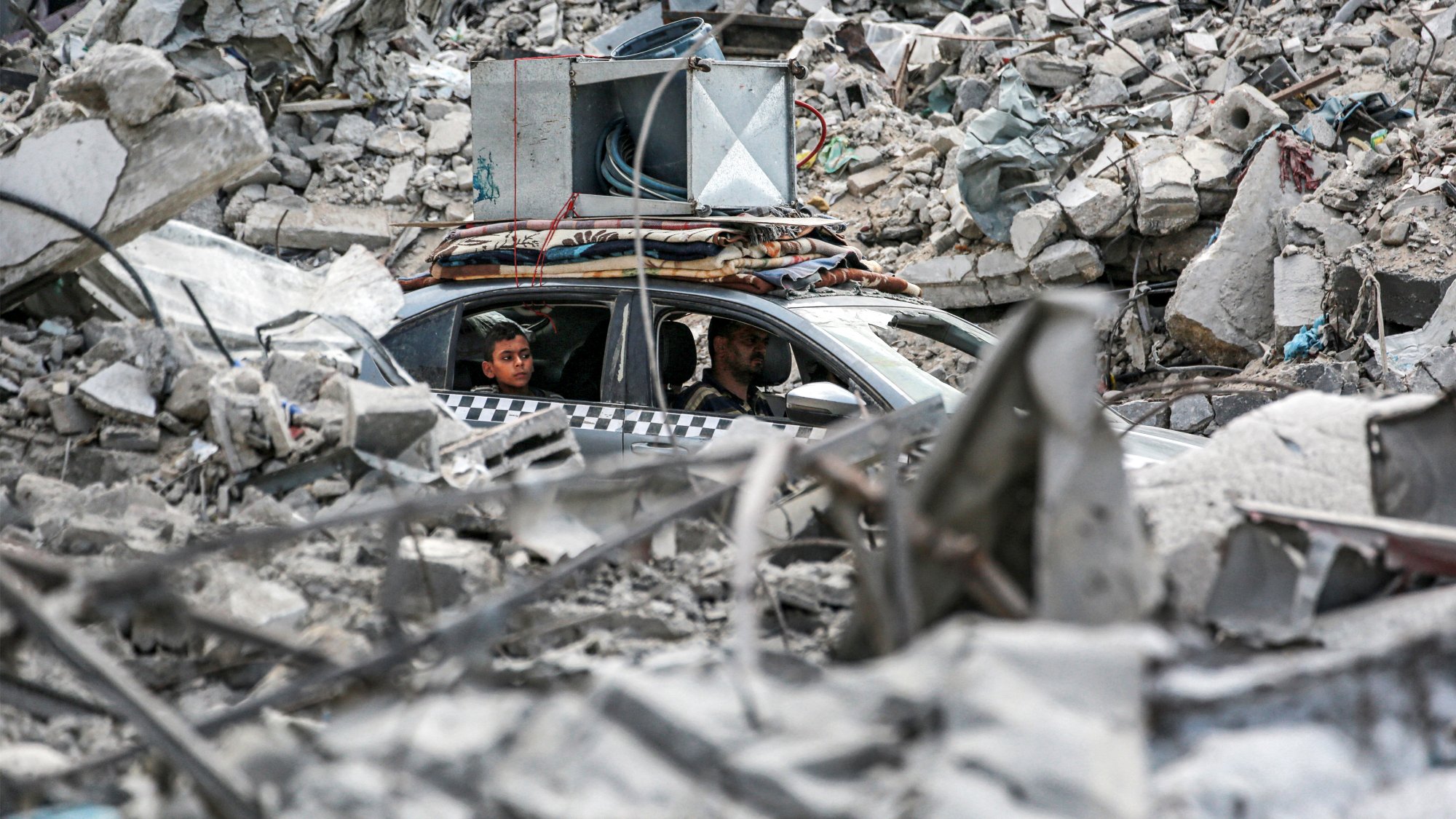 L'image montre un paysage dévasté avec des décombres sur le sol. Au centre, on aperçoit une voiture dans laquelle se trouvent deux personnes. La voiture semble être coincée parmi les débris, qui comprennent des morceaux de béton et d'autres matériaux d'une structure effondrée. L'ambiance générale dégage une impression de destruction et de chaos, témoignant d'une situation difficile.
