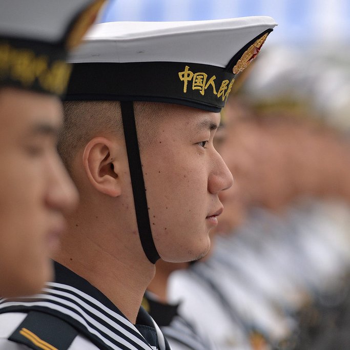 L'image montre des marins en uniforme, probablement lors d'une cérémonie ou d'un événement officiel. On peut voir des hommes alignés, portant des chapeaux distinctifs et des uniformes marins. Les expressions sur leurs visages sont sérieuses, et ils semblent concentrés sur un point donné. L'arrière-plan montre des silhouettes floues d'autres marins, suggérant un grand rassemblement. L'image dégage une ambiance de discipline et de camaraderie militaire.