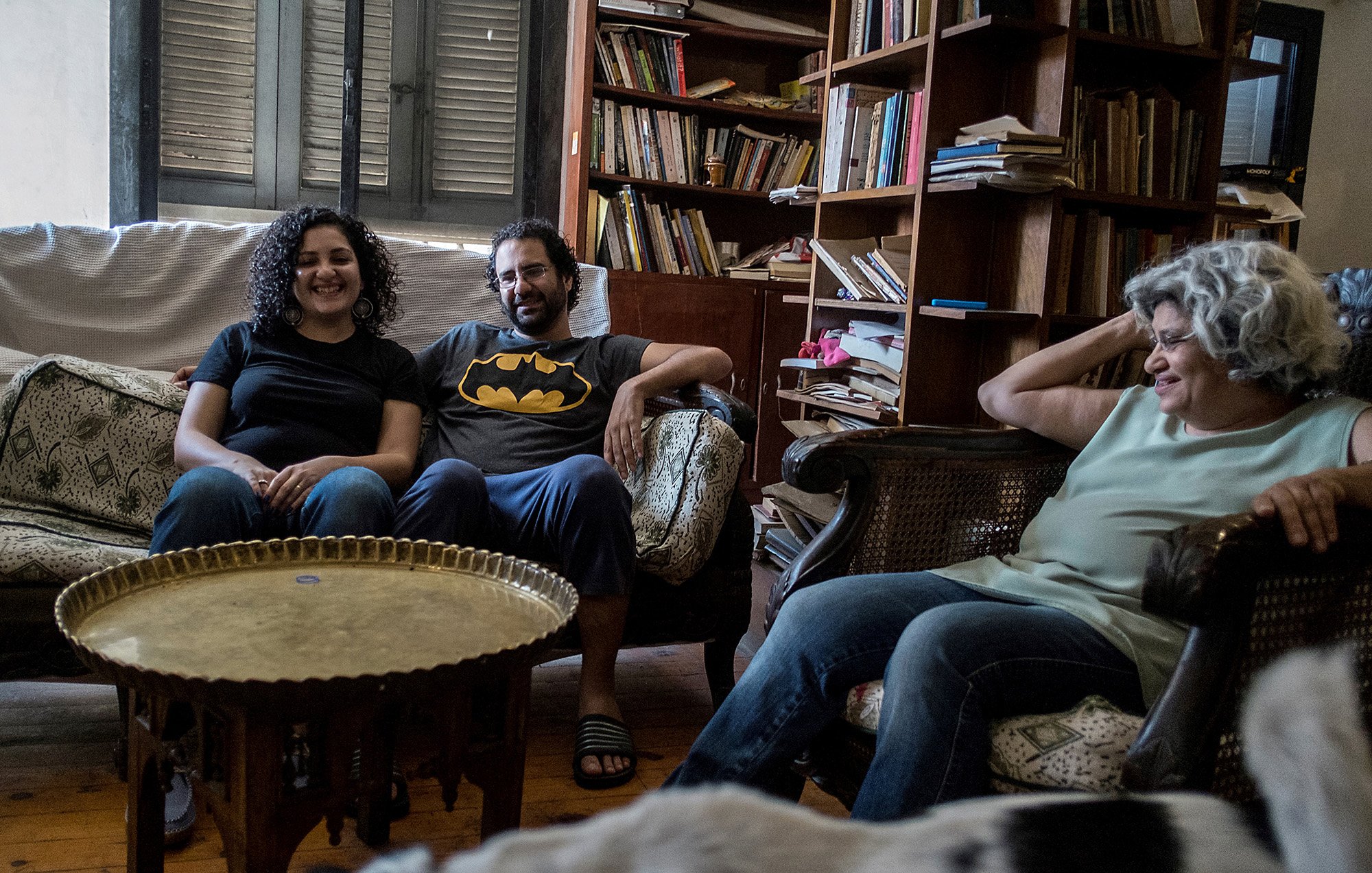 L'immagine mostra un ambiente domestico accogliente, con un soggiorno arredato in modo informale. Ci sono tre persone sedute su poltrone e divani. Due di loro sorridono, mentre un'altra donna, con capelli grigi e ricci, appare rilassata. In mezzo c'è un tavolino rotondo di metallo. Sullo sfondo, ci sono librerie piene di libri, che conferiscono un'atmosfera calda e familiare all'ambiente. Sul pavimento, si intravede un cane.