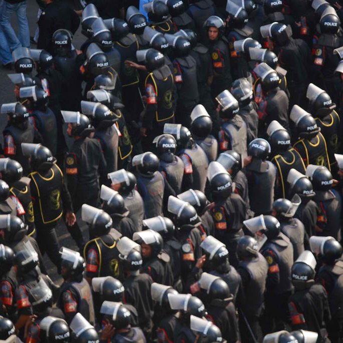 L'image montre un grand groupe de policiers en uniforme, portant des casques et des protections. Ils sont disposés de manière organisée et semblent être en formation, probablement lors d'une manifestation ou d'un événement de sécurité. Les uniformes sont généralement noirs avec des éléments de couleur, et on peut voir des panneaux de protection en plastique. L'atmosphère semble intense et sérieuse, reflétant une situation de gestion de la foule.