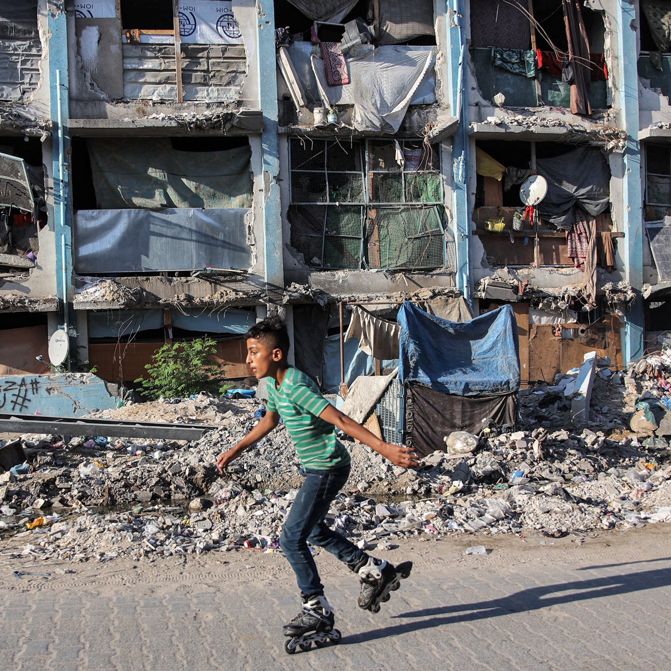 L'image montre un jeune garçon qui skates sur des rollers, se déplaçant avec agilité sur un trottoir. En arrière-plan, des bâtiments en mauvais état sont visibles, avec des fenêtres cassées et des débris éparpillés, ce qui évoque un environnement urbain dégradé. Des morceaux de tissu et d'autres objets sont accrochés aux façades, ajoutant à l'impression générale de désolation. Le contraste entre l'activité du garçon et le décor environnant souligne une certaine résilience face à la pauvreté.