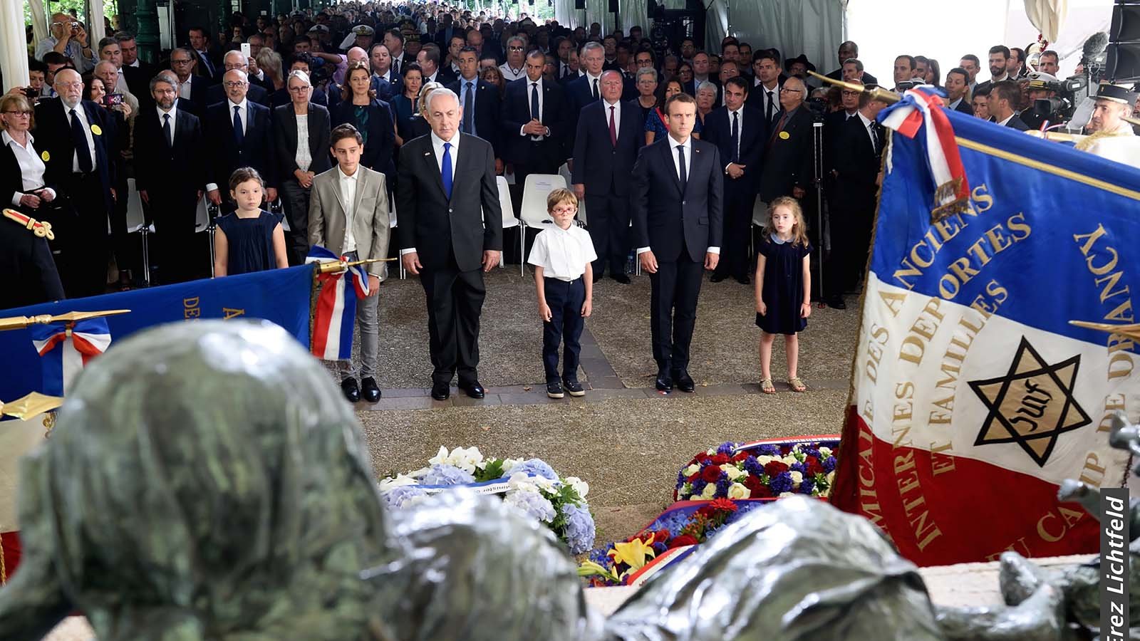 L'image montre une cérémonie commémorative avec un grand nombre de personnes rassemblées sous une tente. On peut voir des adultes en costumes et des enfants bien habillés. Au centre, deux hommes, dont l'un semble être un dirigeant, sont debout, les mains sur le cœur, en signe de respect. Il y a également des drapeaux et des gerbes de fleurs autour d'eux. L'ambiance semble solennelle et respectueuse, avec une attention particulière portée aux éléments de mémoire et d'honneur.