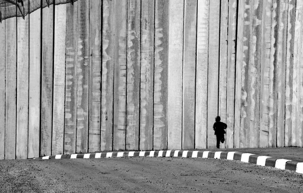 L'image montre un enfant courant le long d'un mur de béton. Le mur est haut et semble imposant, créant une ambiance de séparation ou d'isolement. Le contraste en noir et blanc accentue les textures du mur et la silhouette de l'enfant, qui semble minuscule par rapport à la structure massive. Le sol est désertique avec une route qui tourne légèrement, ajoutant à l'impression de solitude et de mouvement.