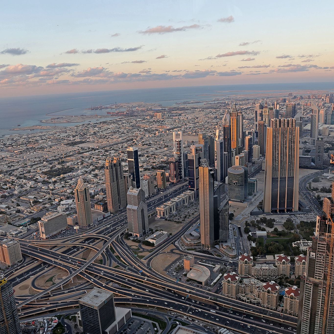 The image depicts a stunning aerial view of a sprawling cityscape, likely a metropolitan area with modern architecture. Skyscrapers dominate the skyline, showcasing a mix of designs and heights. Below, intricate road systems weave through the landscape, indicating a bustling urban environment. In the background, the coastline meets the ocean, with a hint of the horizon showcasing a picturesque sunset. The combination of urban infrastructure and natural beauty creates a striking contrast, highlighting the vibrancy of the city.