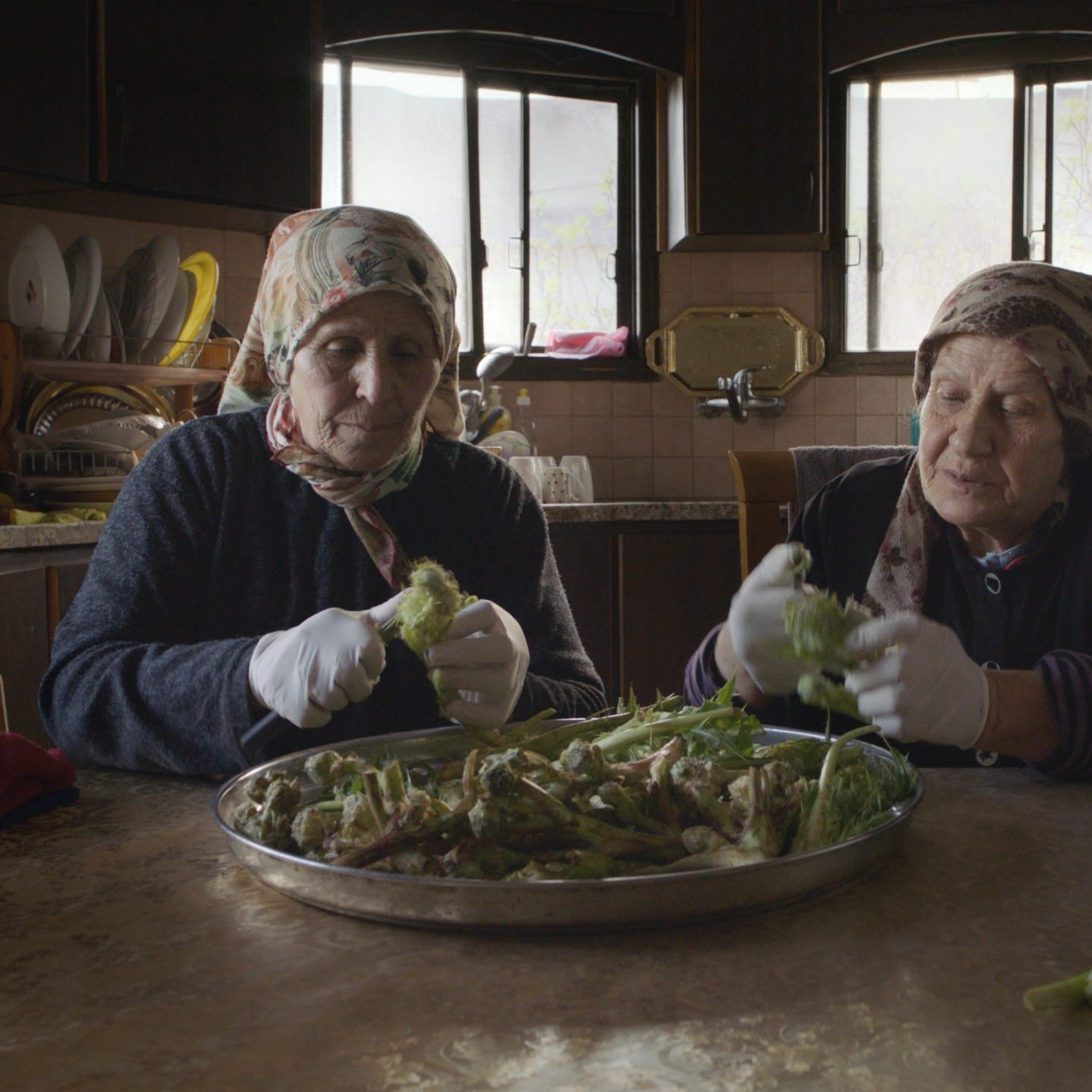 L'image montre deux femmes assises à une table dans une cuisine, en train de préparer des légumes. Elles portent des foulards et des gants blancs. Sur la table, il y a un grand plat rempli de légumes que les femmes épluchent ou nettoient. La cuisine a un décor simple avec des fenêtres qui laissent passer la lumière naturelle. L'atmosphère semble chaleureuse et conviviale, mettant en avant un moment de partage et de tradition culinaire.