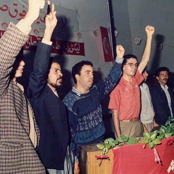 تظهر الصورة مجموعة من الأشخاص واقفين خلف منصة، يرفعون أيديهم في إشارة للتعبير عن الاحتجاج أو الدعم. الخلفية تحتوي على لافتات مكتوبة، مما يشير إلى أن الحدث قد يكون سياسيًا أو اجتماعيًا. الجو العام يبدو حماسيًا، حيث يعكس تفاعل الجمهور مع ما يحدث أمامهم. الأشخاص يرتدون ملابس تتنوع في الألوان والأساليب، مما يعكس حقبة زمنية معينة.