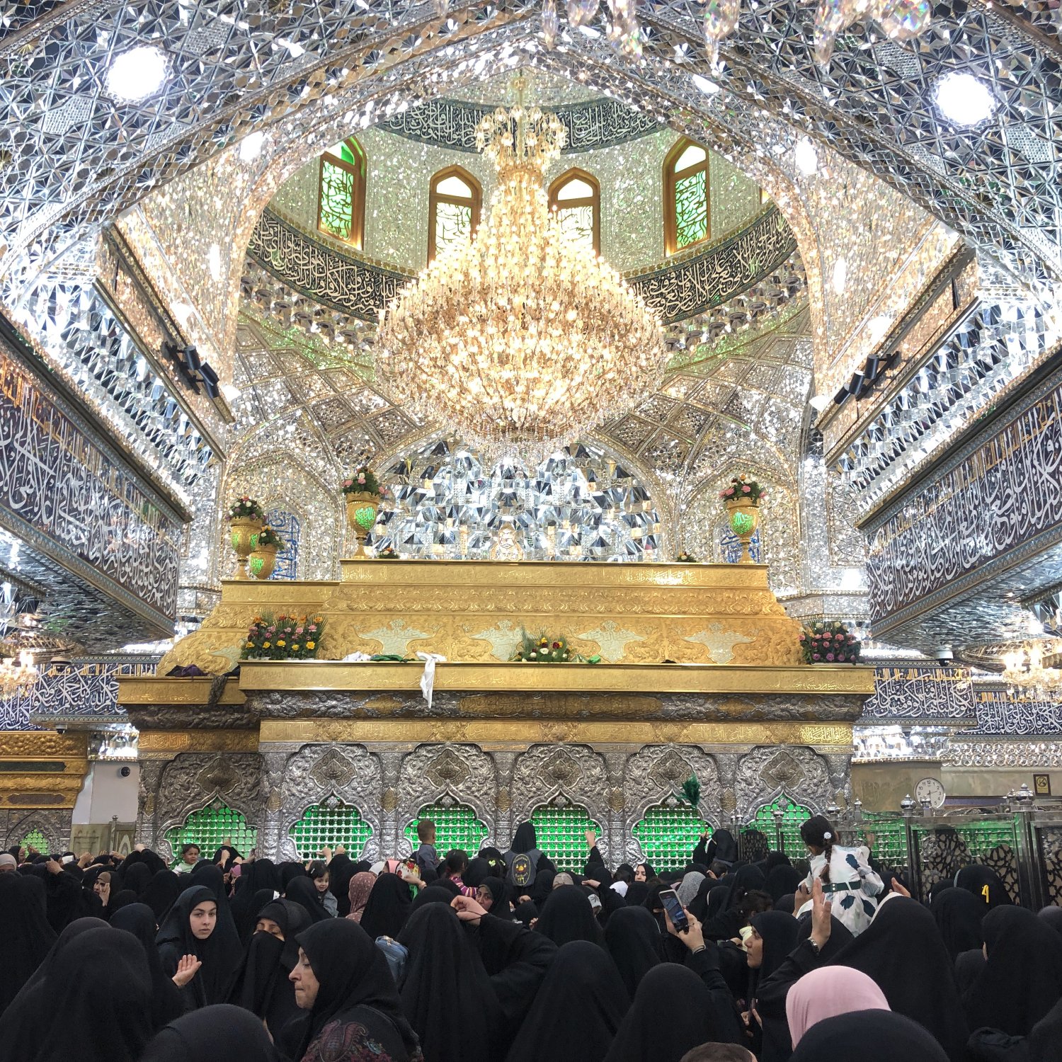 L'image montre un intérieur somptueux, probablement d'un sanctuaire ou d'une mosquée, orné de nombreux détails décoratifs en miroir et de magnifiques motifs. Au centre, on peut voir une structure dorée qui attire l'attention, probablement un mausolée ou une tombe. Le plafond est impressionnant, avec un grand lustre et des fenêtres laissant passer la lumière. Beaucoup de personnes, principalement vêtues de noir, sont rassemblées à l'intérieur, créant une atmosphère de recueillement et de dévotion. Les murs sont également ornés d'inscriptions arabes, ajoutant à la beauté spirituelle du lieu.