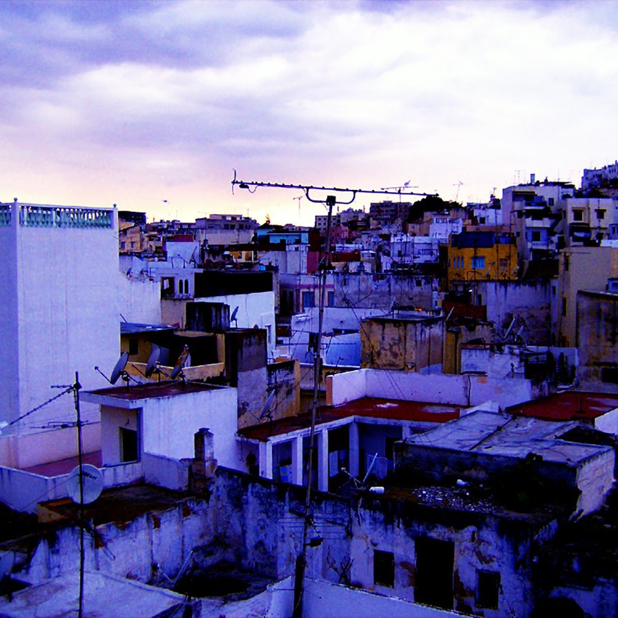 L'image montre une vue urbaine d'un paysage de ville, probablement à la tombée de la nuit. On peut voir plusieurs bâtiments aux structures variées, avec des toits plats et un mélange de couleurs qui évoquent une atmosphère un peu mélancolique. Certains bâtiments semblent légèrement en ruine, avec des murs décrépis et des débris visibles. Le ciel est nuageux, ajoutant une ambiance dramatique à la scène. Des antennes de télévision sont visibles sur les toits, typiques des environnements urbains. L'ensemble donne une impression de vie quotidienne dans une ville qui a du caractère.