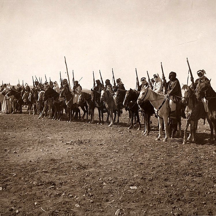 The image depicts a line of mounted warriors or soldiers, likely from a historical context. They are on horseback, holding spears or similar weapons, and are dressed in traditional clothing that suggests their cultural identity. The setting appears to be an open, dry landscape, possibly a desert or arid region. The overall sepia tone gives the photograph an aged appearance, typical of early 20th-century photography. The formation and attire indicate that it could represent a military or ceremonial gathering.