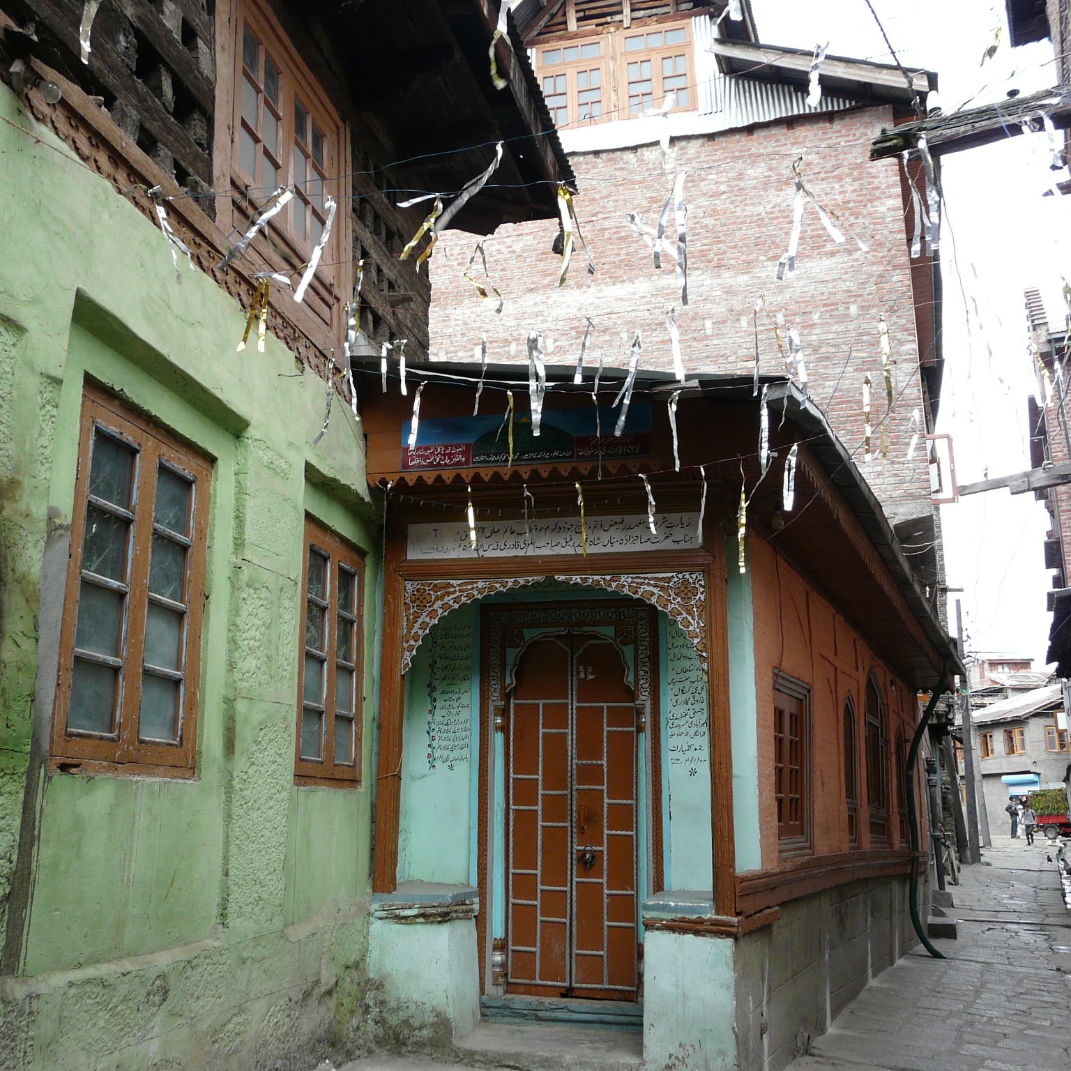 L'image montre une rue étroite et pittoresque dans une vieille ville, avec des maisons en bois et en pierre. On peut voir des façades colorées, notamment des murs verts et oranges, ainsi que des fenêtres à volets en bois. Les bâtiments ont un style architectural traditionnel, avec des détails ornementaux visibles. Des fils sont tendus entre les maisons, probablement pour des décorations ou des lumières. L'atmosphère évoque un sentiment de nostalgie et de charme ancien.