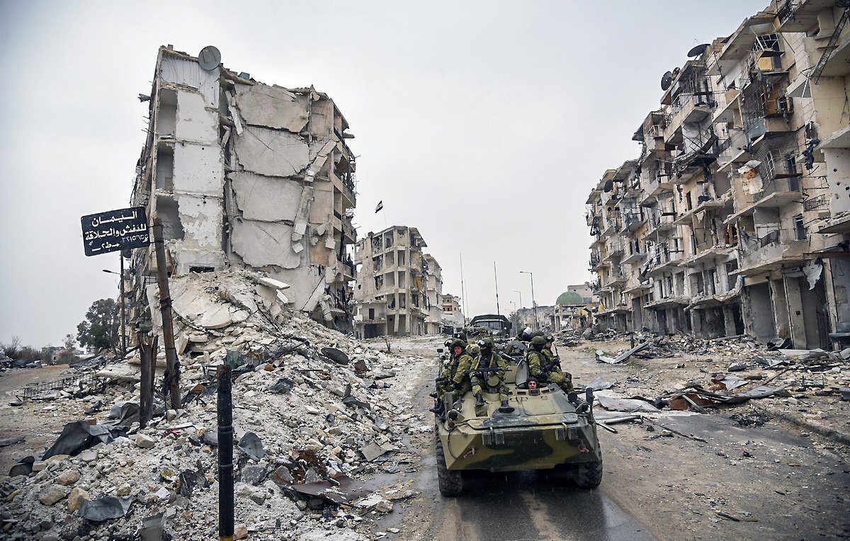 L'image montre une scène de destruction urbaine, probablement à la suite d'un conflit. On peut voir des bâtiments en ruine, avec des façades effondrées et des débris éparpillés sur le sol. Au premier plan, il y a un véhicule militaire, sur lequel des soldats sont présents. L'atmosphère semble sombre et chargée, soulignant les ravages causés par la guerre.
