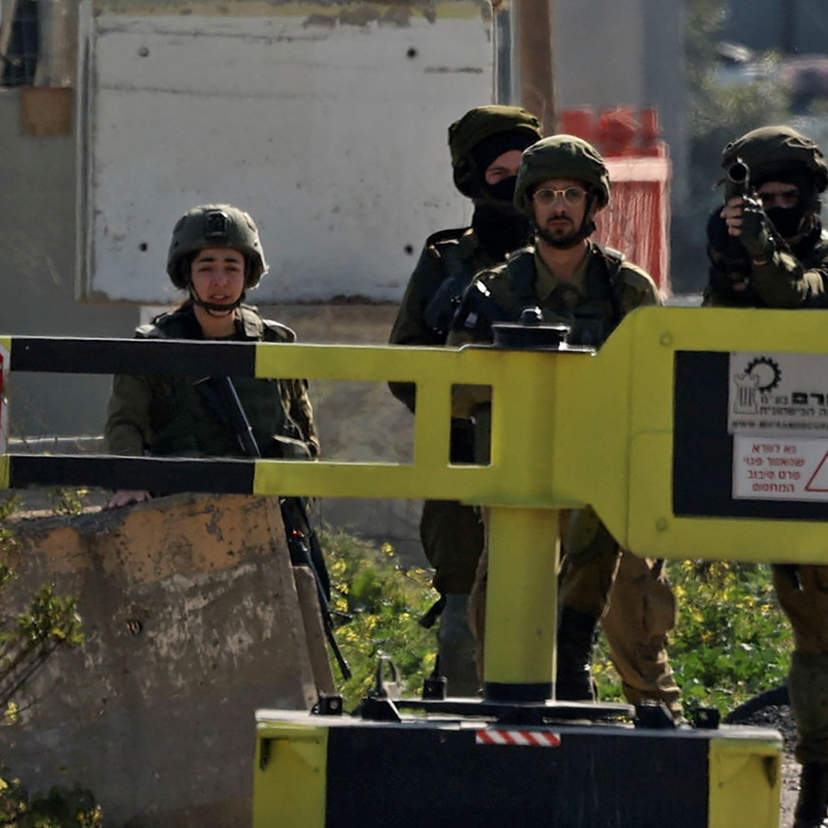 L'image montre des soldats, probablement en situation de contrôle ou de patrouille. Ils portent des uniformes militaires et des équipements de protection. On aperçoit également un panneau de signalisation en forme d'arrêt, qui indique une demande d'arrêt. L'environnement semble être urbain, peut-être dans une zone sensible. Les soldats semblent vigilants et attentifs à leur environnement.