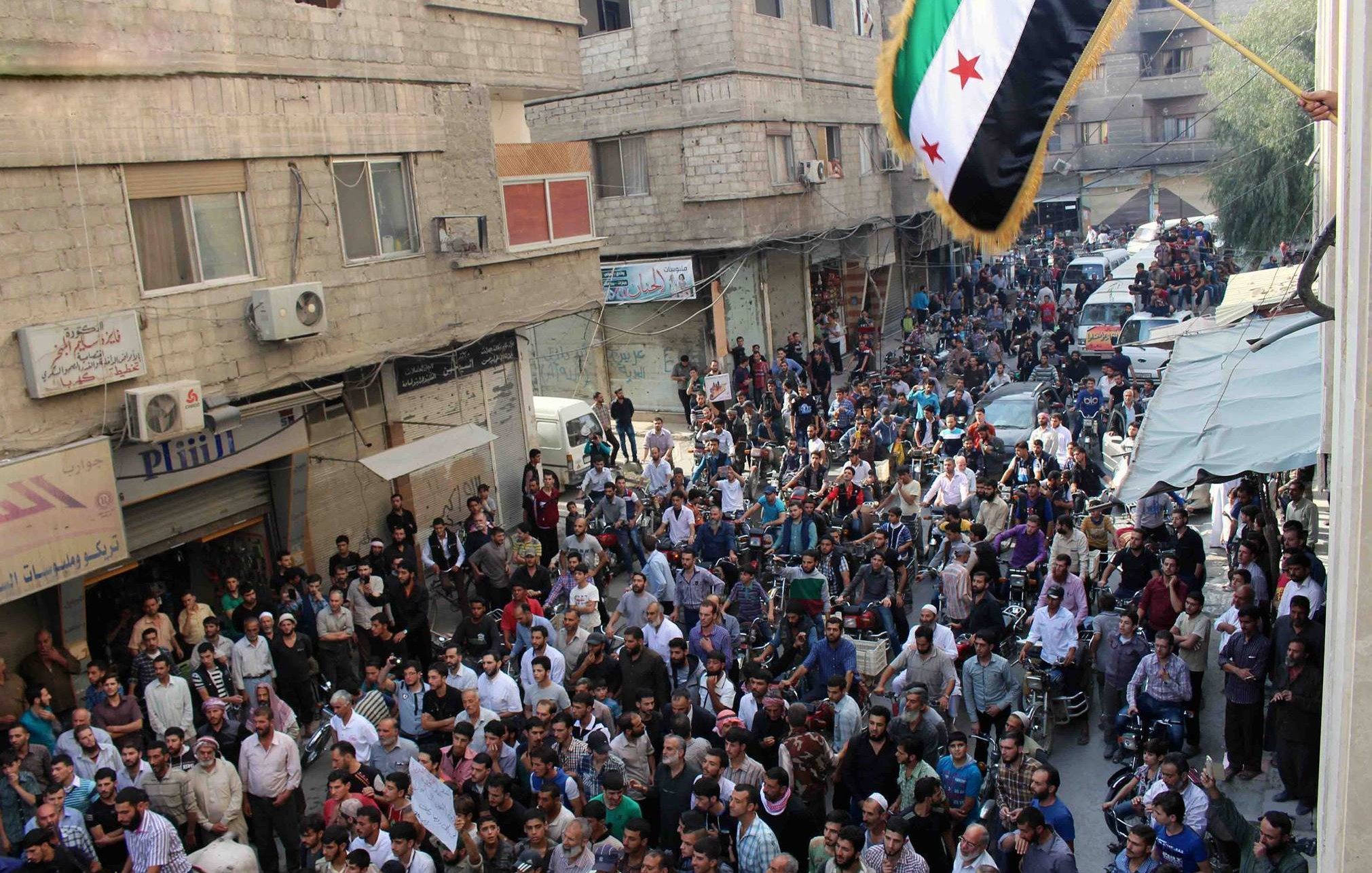 L'image montre une grande foule rassemblée dans une rue, probablement dans une ville syrienne. Les gens semblent se réunir pour une manifestation ou un événement public, avec des pancartes et un drapeau aux couleurs vives, typiquement associé à des mouvements de protestation. Les bâtiments environnants affichent des signes de vie urbaine, et plusieurs véhicules sont stationnés dans la rue. L'atmosphère semble intensément animée, avec une forte concentration de personnes dans l'espace.