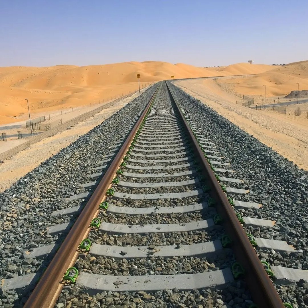 L'image montre une voie de chemin de fer s'étendant à travers un paysage désertique. Les rails, posés sur des traverses en bois, sont entourés de gravier. En arrière-plan, on aperçoit des dunes de sable sous un ciel clair et ensoleillé. Le terrain est principalement aride, et les couleurs dominantes sont des nuances de beige et de gris, ce qui souligne l'isolement et l'immensité de cet environnement désertique.