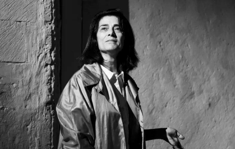 L'image présente une femme qui se tient debout, regardant légèrement vers le haut. Elle a les cheveux longs et foncés, et porte un manteau clair. La lumière joue sur son visage, créant un contraste fort avec l'arrière-plan, qui semble être texturé et neutre. L'ensemble de l'image est en noir et blanc, ce qui accentue l'intensité de l'expression de son visage et la profondeur de la scène.