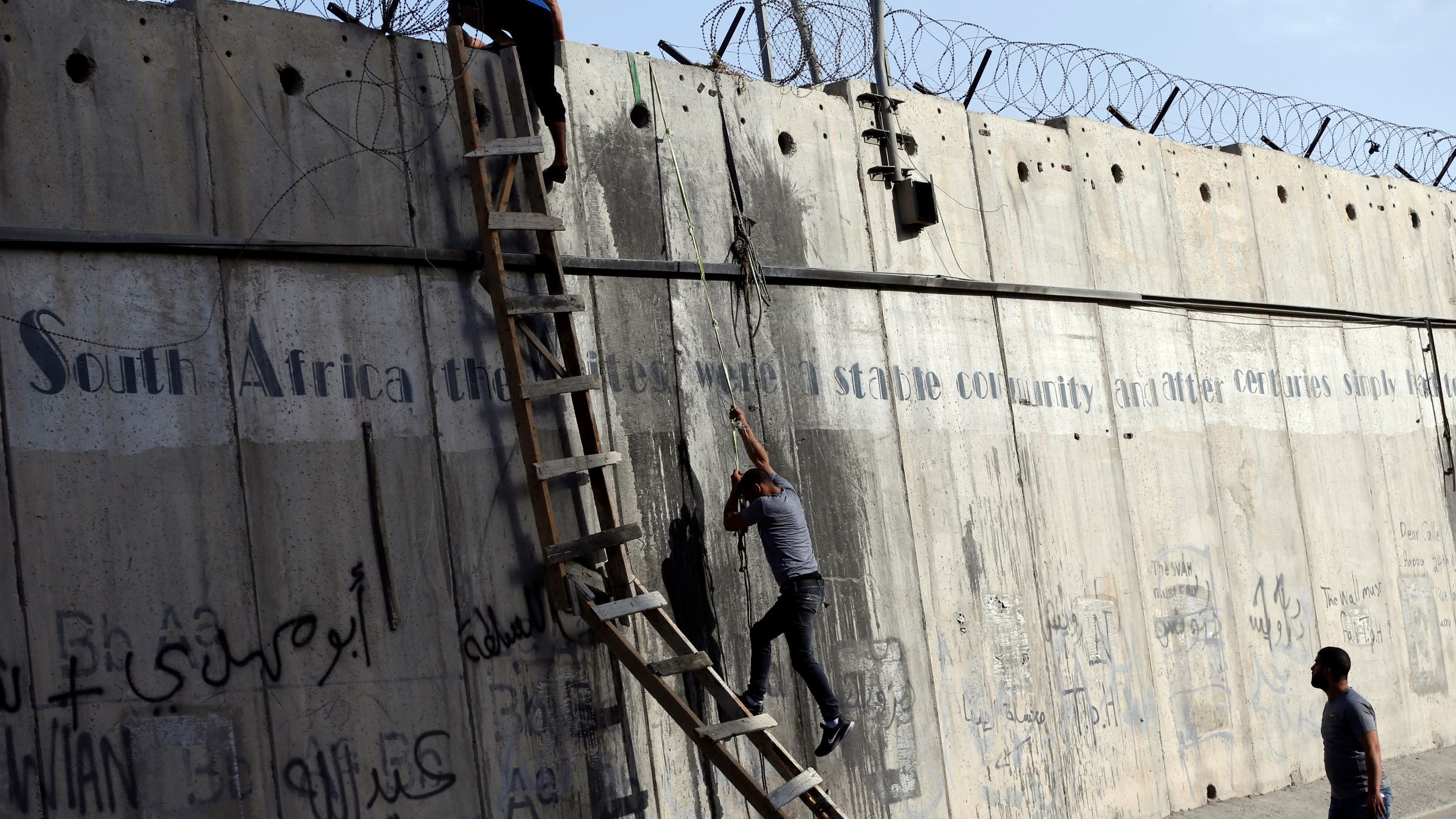 L'immagine mostra due persone che stanno scalando un muro di cemento, sul quale è presente della rete metallica. Dietro di loro, è visibile una scala che qualcuno sta utilizzando per salire. Il muro è coperto da graffiti e scritte, in varie lingue, che sembrano affrontare temi legati alla storia e alla condizione sociale. La scena evoca un senso di tensione e disperazione, con le persone che tentano di superare un ostacolo fisico, simbolo di divisione.