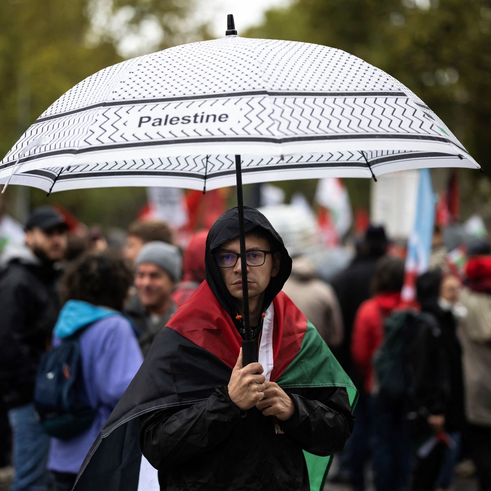 في الصورة، يظهر شخص يحمل مظلة مكتوب عليها "فلسطين"، ويقف وسط حشود من الناس في تظاهرة. يرتدي الشخص علم فلسطين حول كتفيه، وتبدو بعض وجوه المتظاهرين في الخلفية، مع تأثيرات الطقس الممطر، حيث يحمل البعض مظلات أخرى. تعكس الصورة أجواء التظاهر والدعم للقضية الفلسطينية.