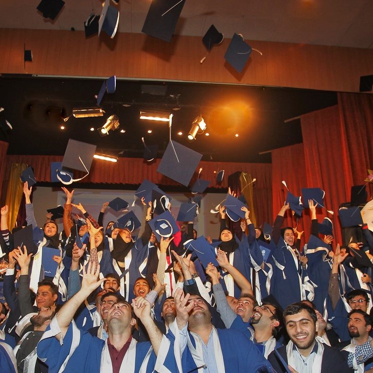 L'image montre une célébration de remise de diplômes. De nombreux étudiants, vêtus de toges et de mortiers de graduation, se tiennent ensemble sur scène. Ils lèvent leurs chapeaux en l'air, exprimant leur joie et leur excitation. L'atmosphère est festive, avec des sourires sur les visages des diplômés, soulignant un moment marquant de leur parcours académique. Des lumières et des décorations de scène ajoutent à l'ambiance festive.