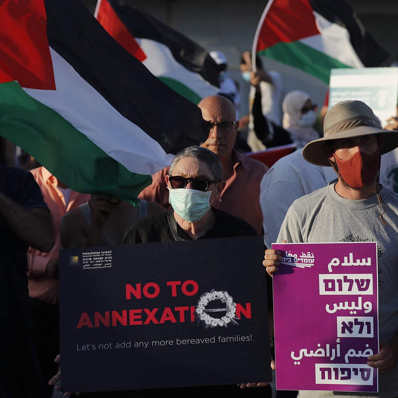 L'image montre une manifestation avec des participants portant des masques. Plusieurs personnes tiennent des drapeaux palestiniens. Des pancartes sont visibles, certaines affichant des messages tels que "NO TO ANNEXATION" (Non à l'annexion) et d'autres slogans en arabe et en hébreu. L'ambiance semble être celle d'une mobilisation pour une cause sociale ou politique, avec un engagement visible des manifestants.