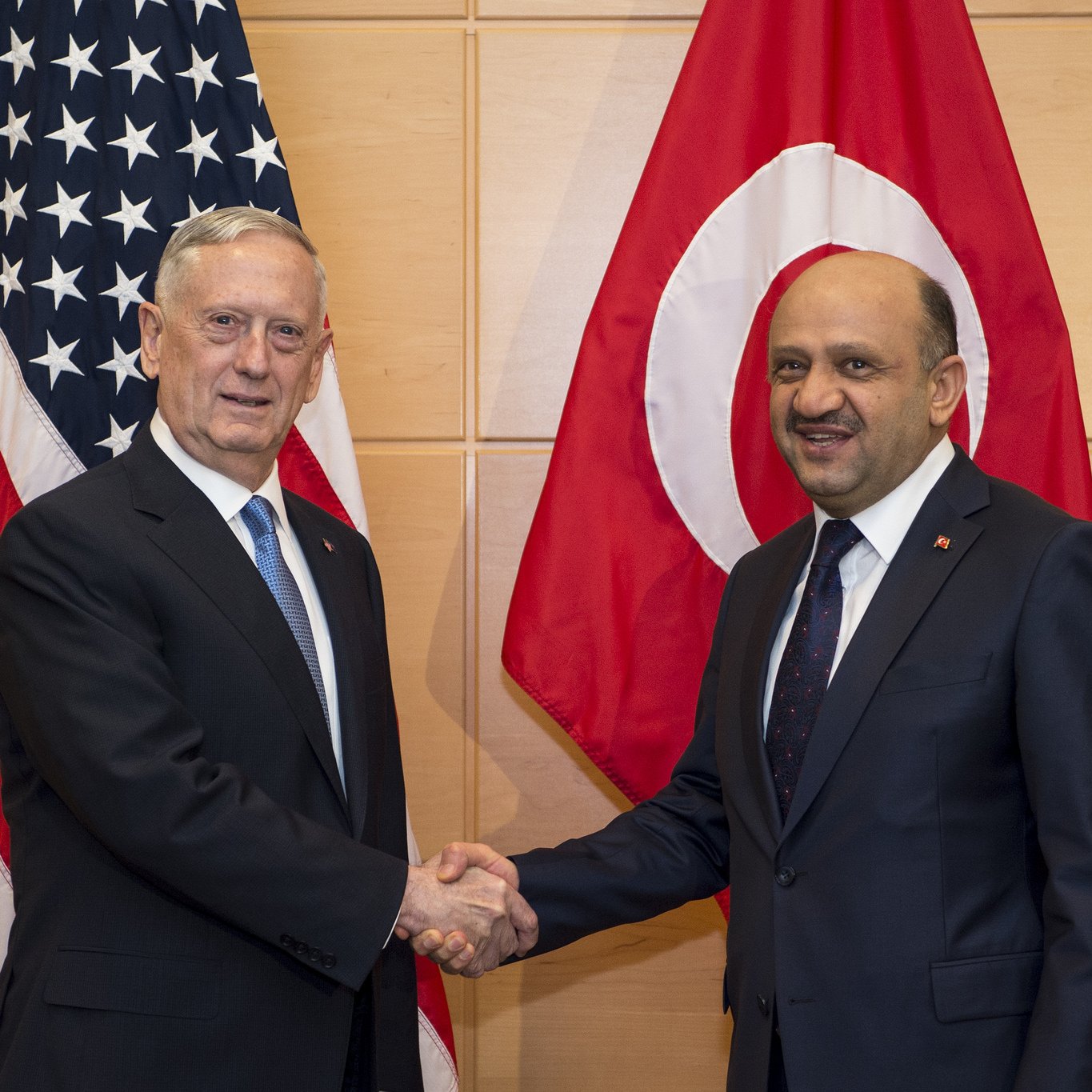 L'image montre deux hommes se serrant la main, probablement en signe de coopération ou d'accord. À l'arrière-plan, on peut voir deux drapeaux : celui des États-Unis à gauche et celui de la Turquie à droite. Les deux hommes portent des costumes formels, et l'ambiance semble être celle d'une rencontre officielle.