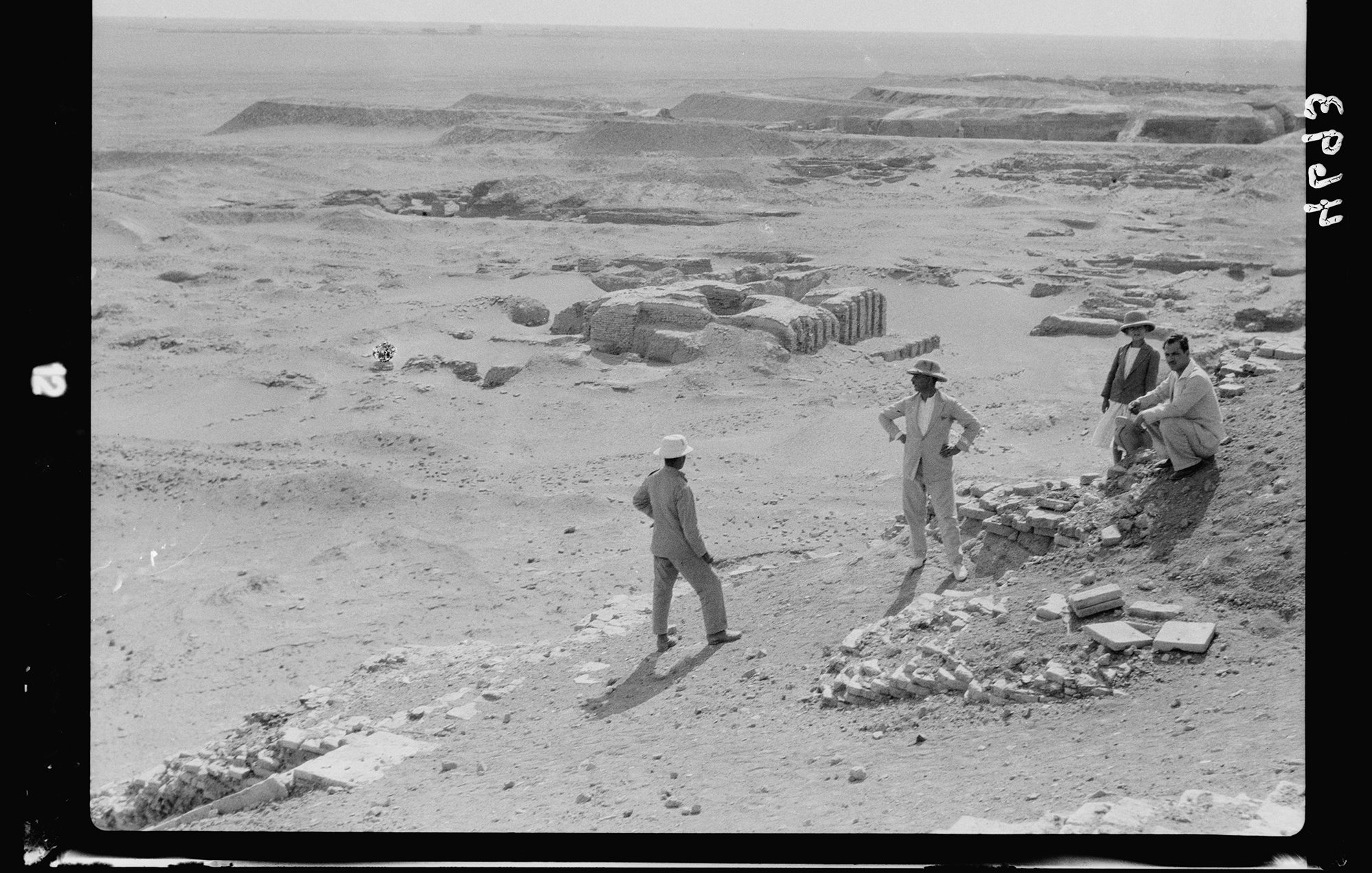 L'image montre une scène archéologique dans un paysage désertique. Plusieurs hommes, habillés de manière décontractée et portant des chapeaux, sont visibles. Ils semblent discuter ou examiner des fragments de pierres et des structures en ruines au sol. En arrière-plan, on peut voir des sites de fouille avec des vestiges qui suggèrent une activité archéologique en cours. L'environnement est aride, et le ciel paraît clair, évoquant une ambiance de travail sur un site ancien.
