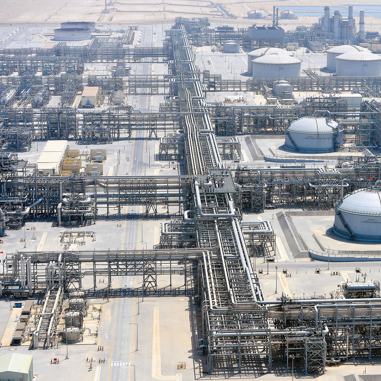 The image depicts a large industrial facility, likely an oil refinery or petrochemical plant. It features numerous large storage tanks, interconnected pipelines, and extensive infrastructure. The site is expansive, suggesting significant industrial activity, and it appears to be located in a relatively arid region, as indicated by the surrounding landscape. There are various structures for processing and storage, emphasizing the complexity and scale of the operation.