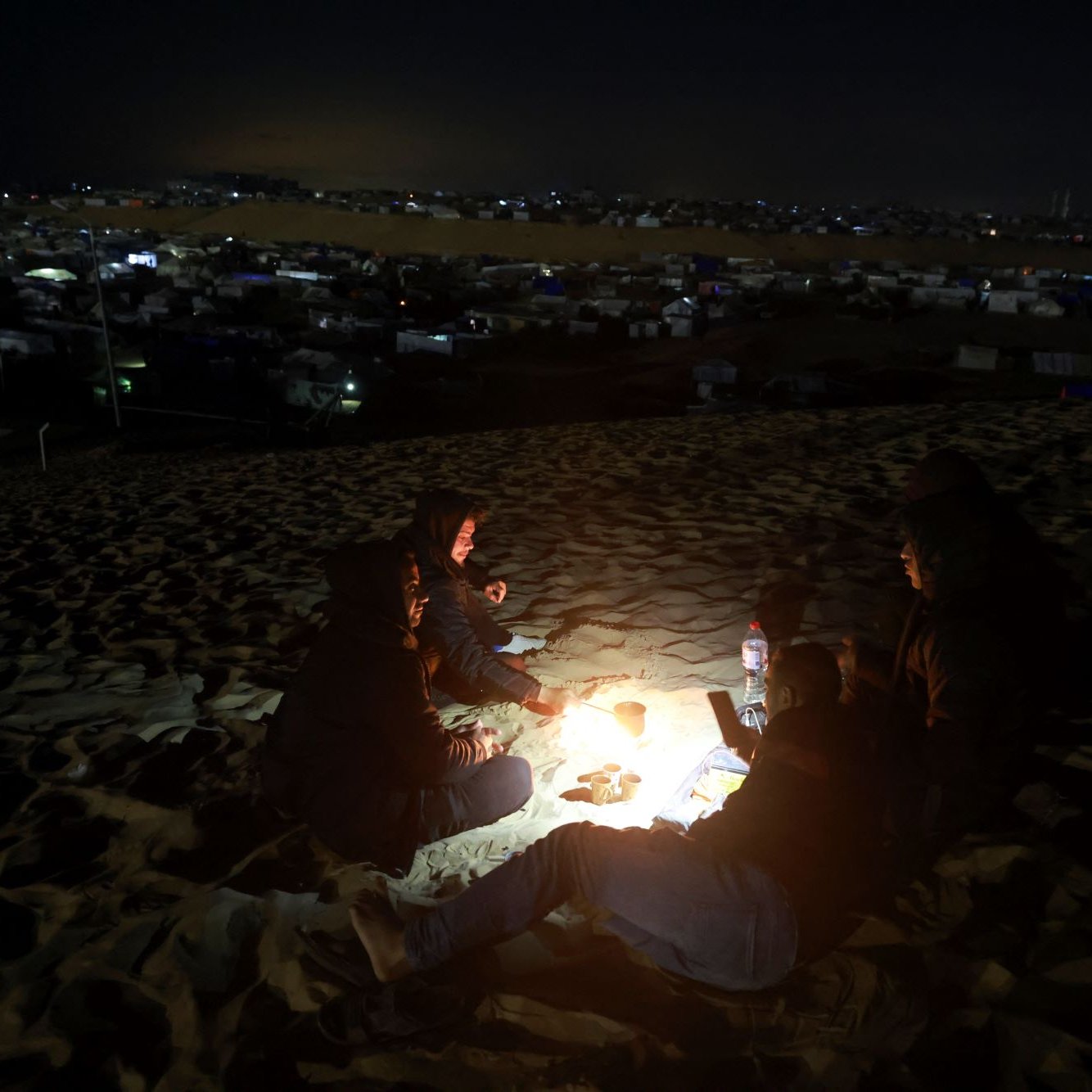 L'image montre un groupe de personnes assises autour d'un feu de camp sur une dune de sable, en pleine nuit. Ils semblent se réchauffer et passer un moment ensemble. En arrière-plan, on peut apercevoir des tentes et d'autres structures, suggérant qu'ils se trouvent dans un campement. La lumière du feu éclaire leurs visages et crée une atmosphère chaleureuse malgré l'obscurité environnante.